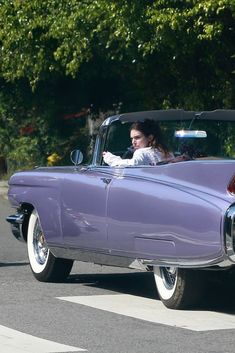 a woman in a purple convertible car driving down the street with trees in the background