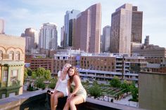 two young women sitting on the ledge of a building in front of cityscape