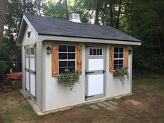 a small shed with two windows and shutters on the doors is shown in front of some trees