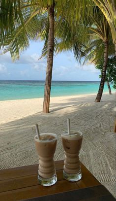 two drinks sitting on top of a wooden table next to the ocean and palm trees