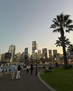 people are walking along the waterfront in front of some tall buildings and palm trees at dusk