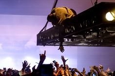 a crowd of people at a concert with their hands in the air and one person on a platform above them