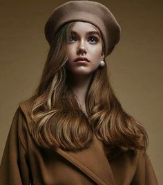 a woman in a brown coat and hat with long hair wearing a white pearl earrings