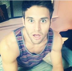 a man in tank top laying on bed looking at the camera with surprised look on his face