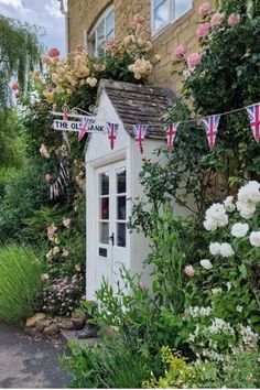 a small white shed with flags hanging from it's roof next to flowers and bushes