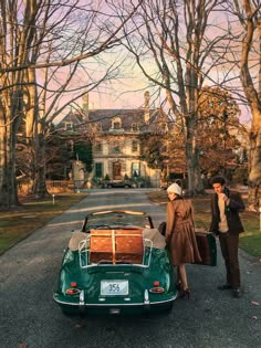 a man and woman standing next to a green car with luggage on the back in front of a large house