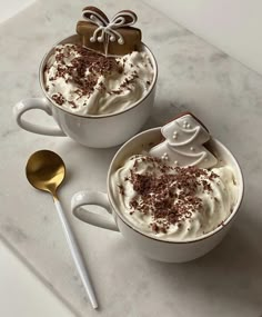 two white mugs filled with whipped cream and topped with chocolate, sprinkles and a bow