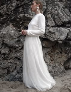 a woman wearing a white dress standing in front of some rocks and looking off into the distance