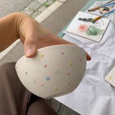 a person holding a white bowl with flowers on it and painting supplies in the background