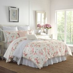 a white bed with pink flowers on the comforter and pillows in front of a window