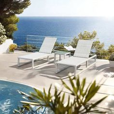 two white lounge chairs sitting next to a swimming pool near the ocean on a sunny day