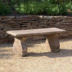 a stone bench sitting in front of a rock wall and graveled area with plants