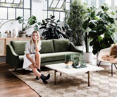 a woman sitting on top of a green couch in a living room next to potted plants