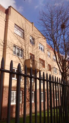 an apartment building behind a black fence with trees in the foreground and a bird flying overhead