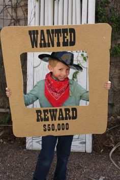 a young boy wearing a cowboy hat and scarf holding up a cardboard wanted reward sign