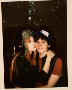 two people that are kissing each other in front of some trees at night with one person wearing a hat