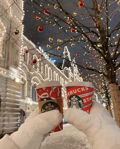 two cups of starbucks coffee in the snow with christmas lights on the trees behind them