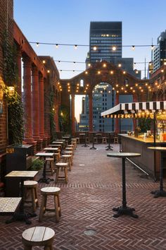 an outdoor patio with tables and chairs, lights strung from the ceiling, and brick buildings in the background