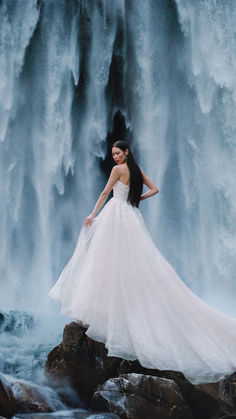 a woman in a wedding dress standing on rocks near a waterfall