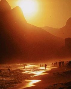 people are walking on the beach as the sun goes down in the mountains behind them