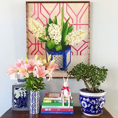 flowers and books on a table in front of a wall hanging with an art piece