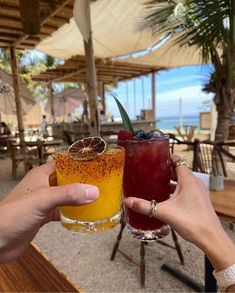 two people holding up glasses with drinks in front of an ocean and beach side restaurant