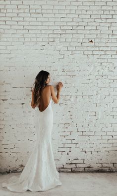 a woman standing in front of a brick wall wearing a white wedding dress with open back