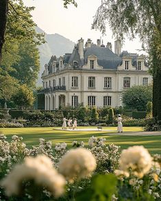 a large white house surrounded by lush green trees and flowers in the foreground with people walking around