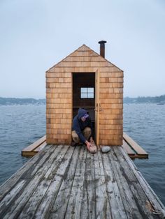 a man kneeling down in front of a small wooden cabin on top of a body of water