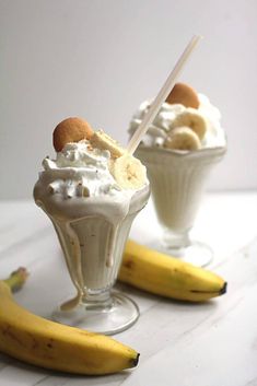 two glasses filled with ice cream and banana slices on a white table next to some bananas