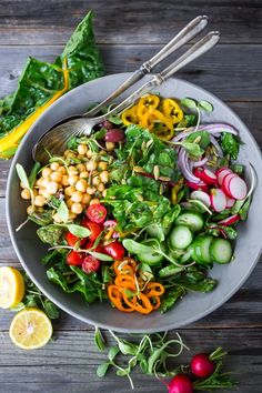 a bowl filled with salad and vegetables on top of a wooden table next to lemon wedges