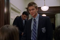 a young man in a suit and tie standing in front of a mirror with other people looking at him