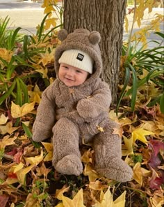 a baby in a teddy bear costume sitting under a tree with leaves on the ground