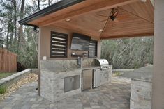 an outdoor kitchen with grill and sink under a wooden roof over a brick patio area