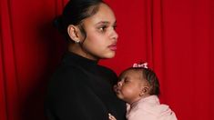 a woman holding a baby in front of a red curtain with her hands on her chest