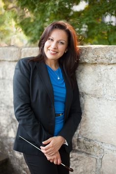 a woman leaning against a stone wall wearing a blue shirt and black blazer with her hands in her pockets