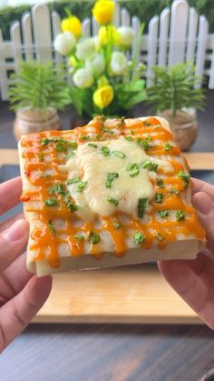 someone is holding up a piece of food with cheese and sauce on it in front of some flowers