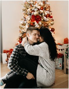 a man and woman sitting in front of a christmas tree with their arms around each other