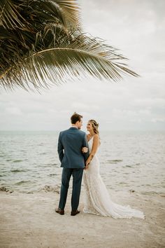 a bride and groom standing on the beach under a palm tree