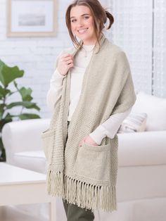 a woman standing in front of a couch wearing a white sweater and fringed shawl