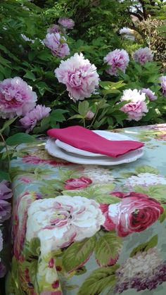 a table covered with pink flowers in the middle of green bushes and trees behind it