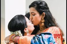 two women are kissing each other in front of a white wall and one is wearing an orange sari