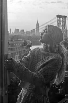 black and white photograph of woman looking out window at cityscape with bridge in background