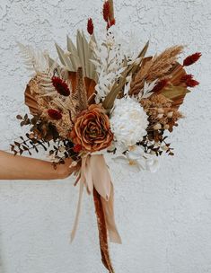 a person holding a bouquet with flowers and leaves on the outside of their arm, in front of a white wall