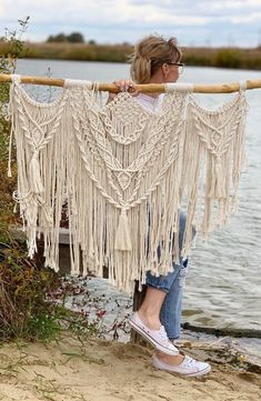 a woman standing on the beach holding up a macrame wall hanging from a wooden pole