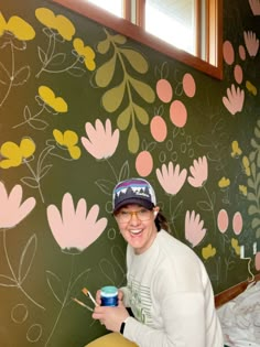 a woman sitting on a bed holding a paintbrush and painting the wall with flowers