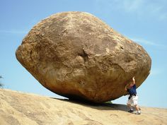 a man standing next to a large rock on top of a sandy hill under a blue sky