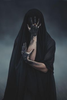 a woman with her hands painted black and wearing a hooded outfit, holding two outstretched hands in front of her face