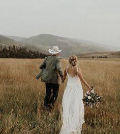 a bride and groom holding hands walking through tall grass with mountains in the back ground