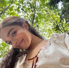 a woman with long hair wearing a white dress and smiling at the camera while standing in front of trees